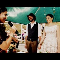 marina bastos celebrando casamento com microfone na mão, sorrindo e olhando para os noivos, que estão sorrindo e de mãos dadas